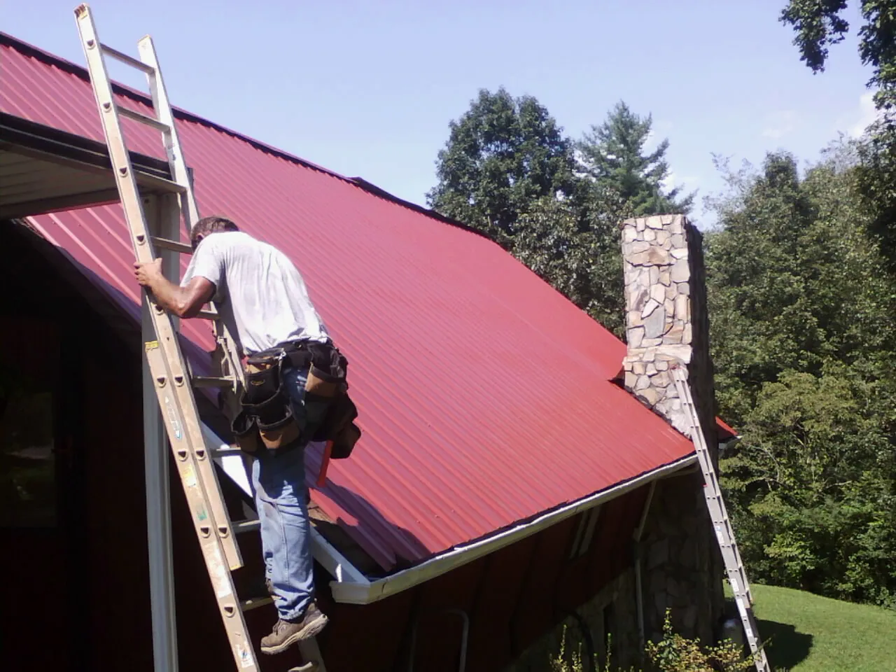 Roofer on a ladder beside a red metal roof and stone chimney — Soffit & Fascia Repair by Roofing Brothers in Glade Hill